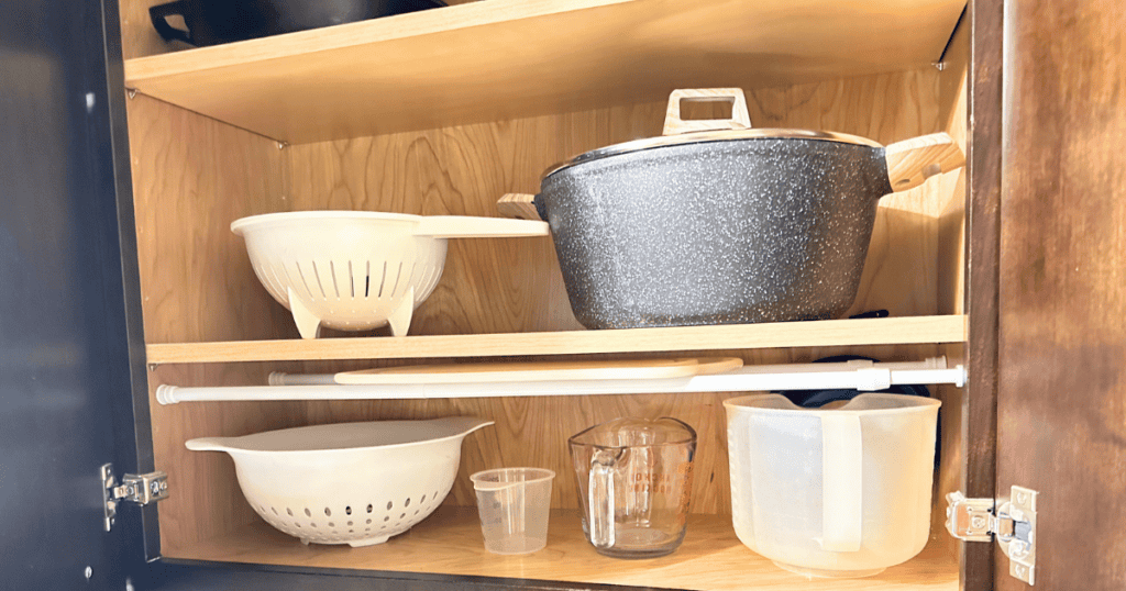 tension rod in a cabinet holding a cutting board over top of measuring cups