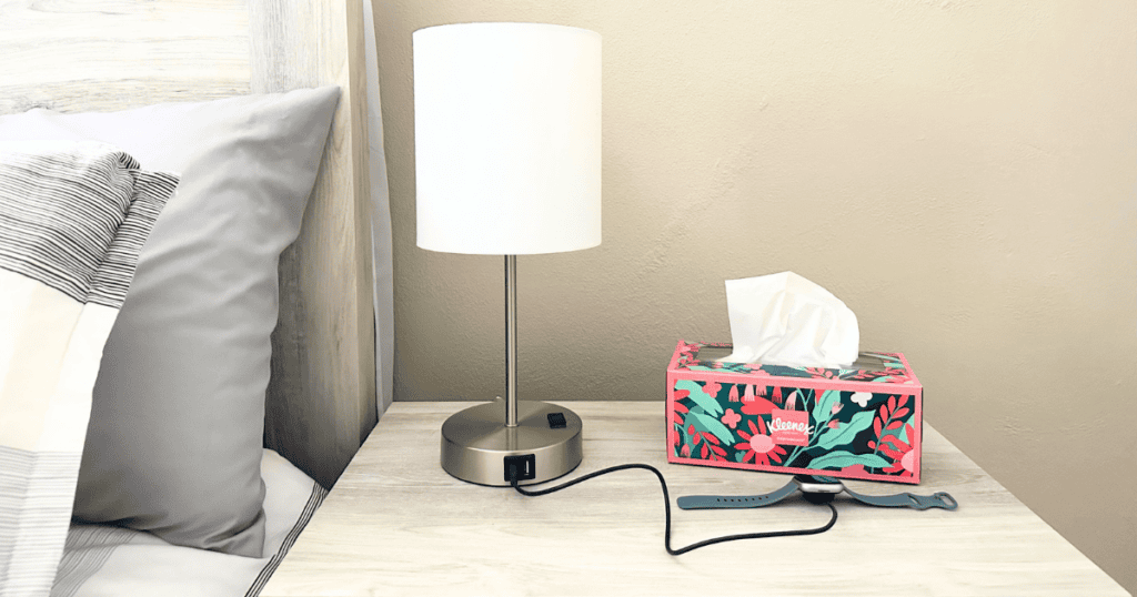 A lamp with two usb charging ports and a watch charging in one of them. A nightstand beside a bed