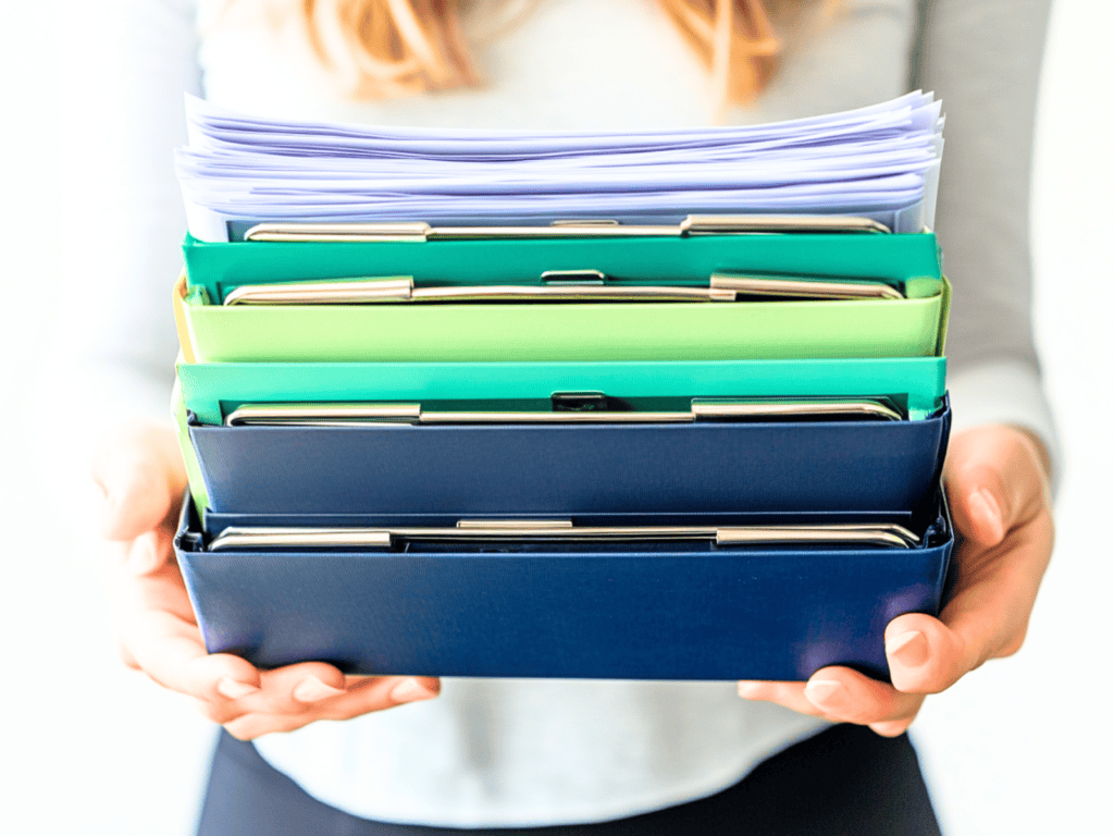 A woman holding four binders and a stack of papers