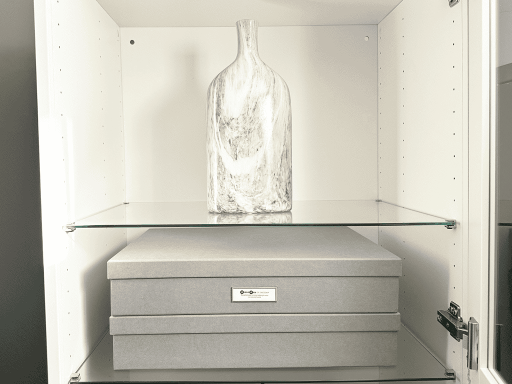 Two gray storage boxes sitting on a shelf below a vase in my spare bedroom.