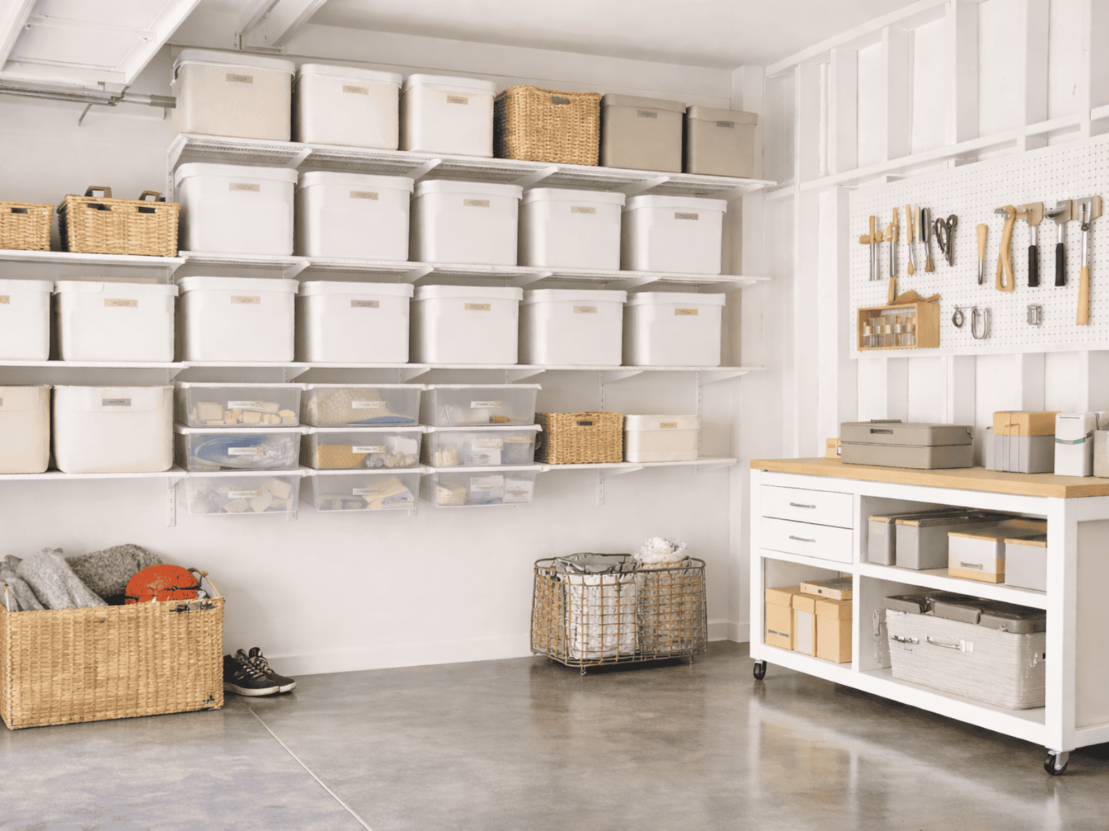 Bright, organized garage with white wall-mounted shelves, labeled storage bins, woven baskets, a pegboard tool wall, and a rolling workbench on a polished concrete floor.
