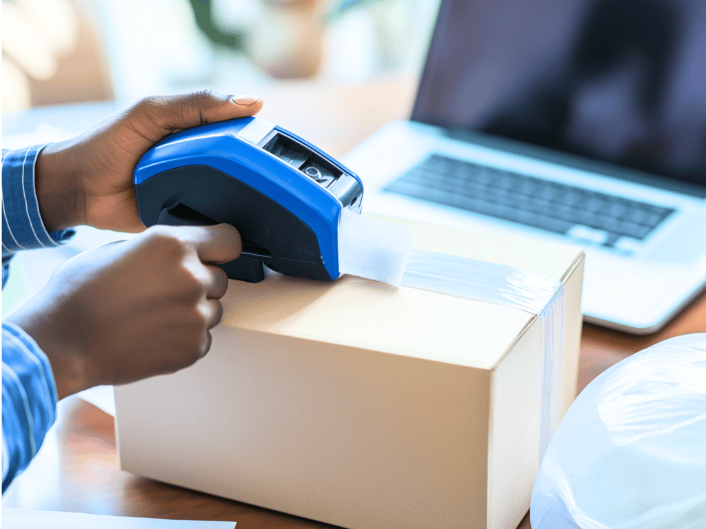 A person taping a box closed with a laptop sitting in the background.