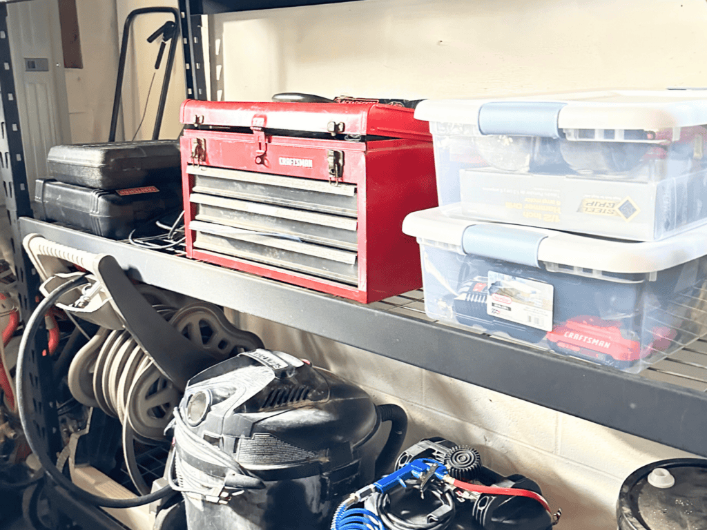 A tool box and plastic totes on a black wire shelf in a garage.