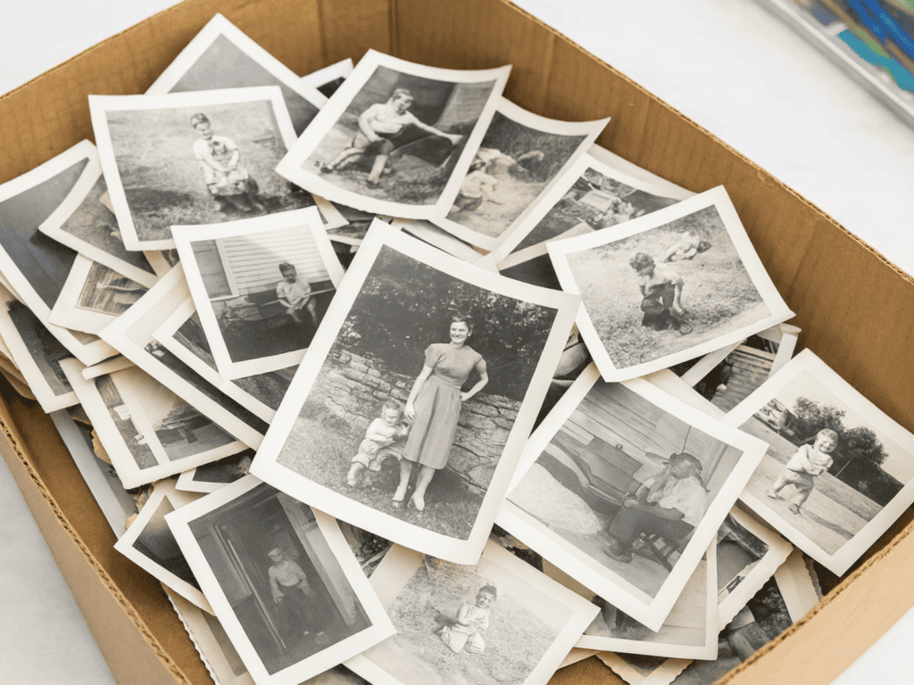 A cardboard box filled with vintage black-and-white photographs, showing various family moments and childhood scenes scattered loosely inside the box.