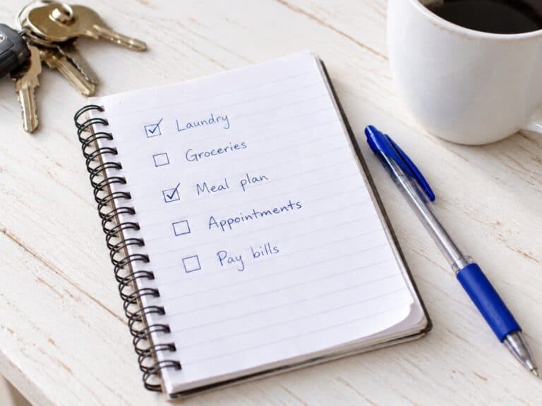 Handwritten organization checklist in a spiral notebook on a white table beside a blue pen, coffee mug, and keys.