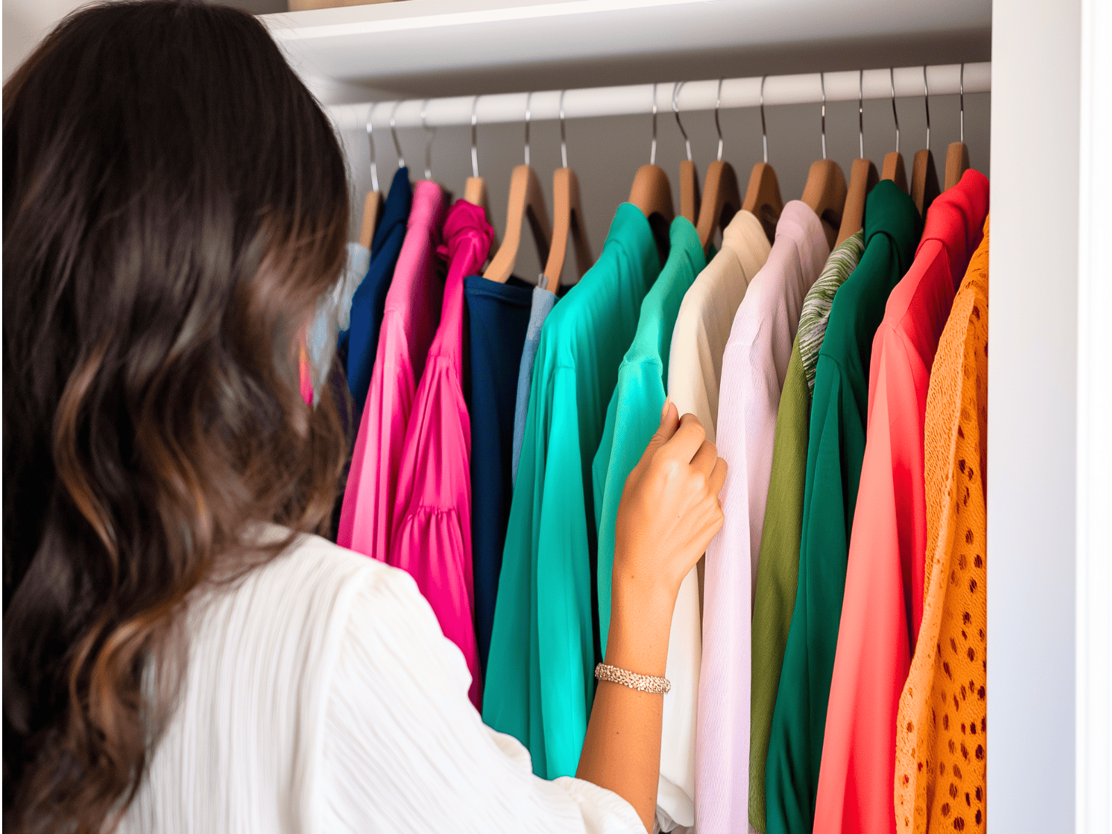 A woman facing a wardrobe filled with colorful clothes on hangers, with her hand reaching towards a teal blouse.