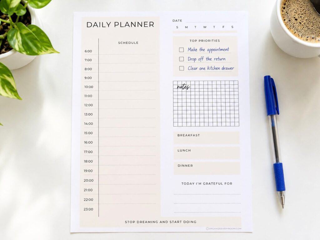 Daily planner with three handwritten priorities on a bright white table beside a blue pen, coffee mug, and green plant.