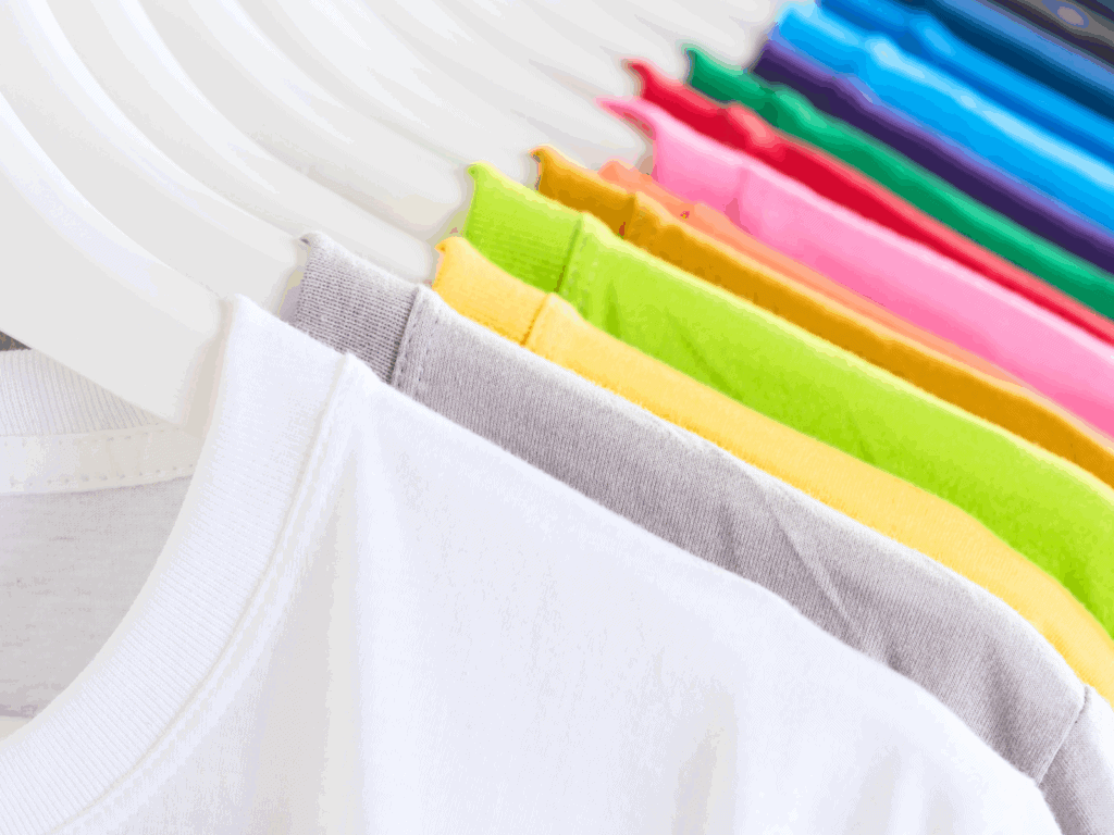 A close-up view of colorful t-shirts neatly hanging on white hangers. The shirts are arranged in a gradient, starting with white, gray, yellow, green, and progressing through pink, red, purple, blue, and teal, creating a vibrant and organized display.