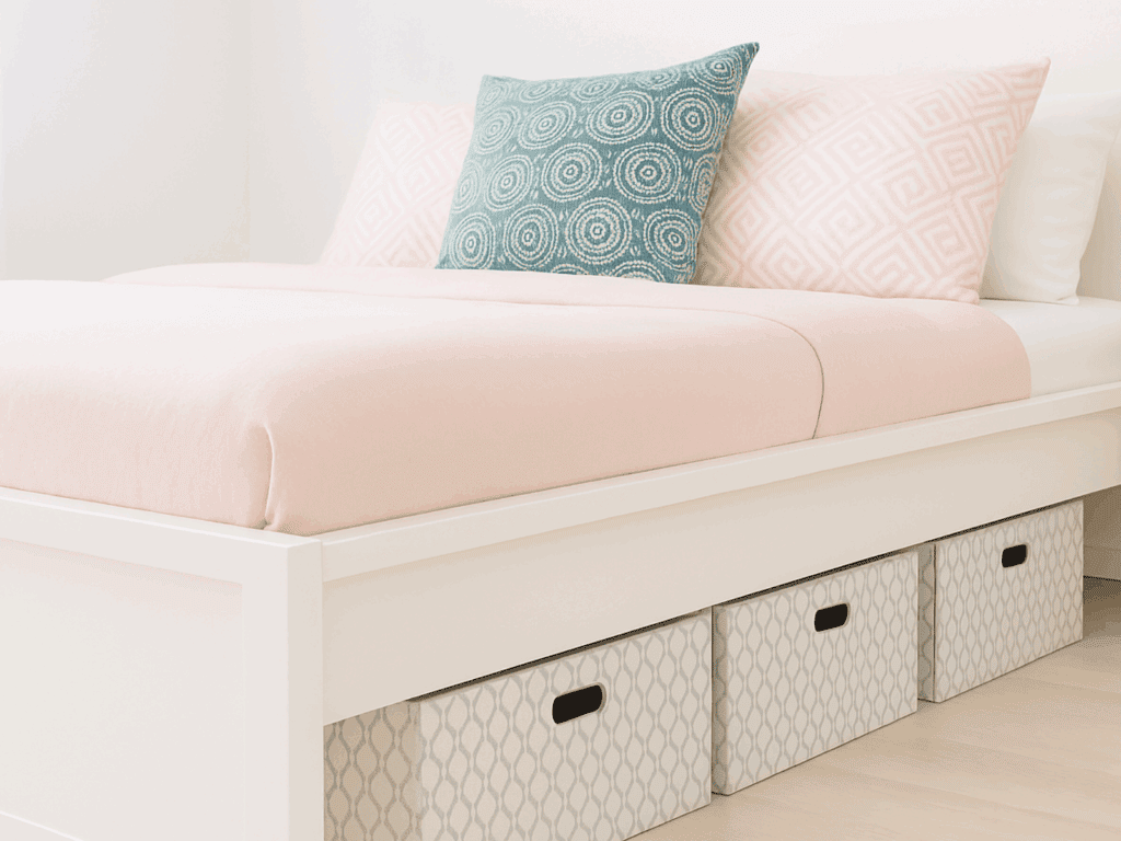 Bright and airy bedroom featuring a white platform bed with soft pink bedding, a white pillow, a pink and white geometric pillow, and three patterned storage bins neatly placed underneath.