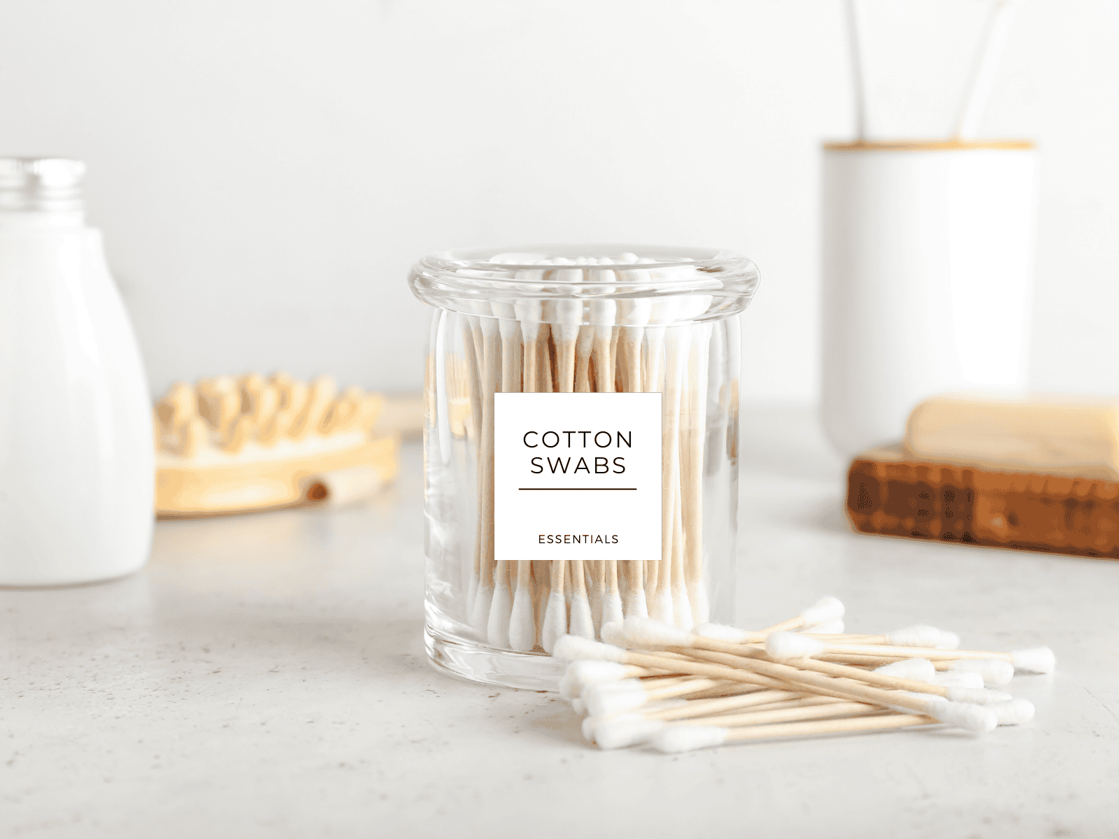 A clear glass jar labeled “Cotton Swabs – Essentials” sits on a clean bathroom counter with wooden cotton swabs inside.