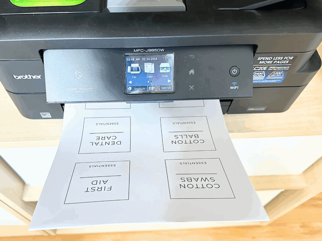 A Brother Work Smart Series printer printing a sheet of black-and-white labels. The labels are neatly designed and include titles such as "COTTON BALLS," "DENTAL CARE," "FIRST AID," and "COTTON SWABS," each under the heading "ESSENTIALS."