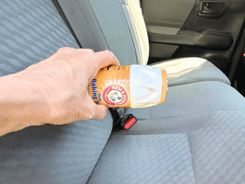 Hand sprinkling baking soda onto a car seat to deodorize and freshen the fabric as part of a DIY car cleaning hack.