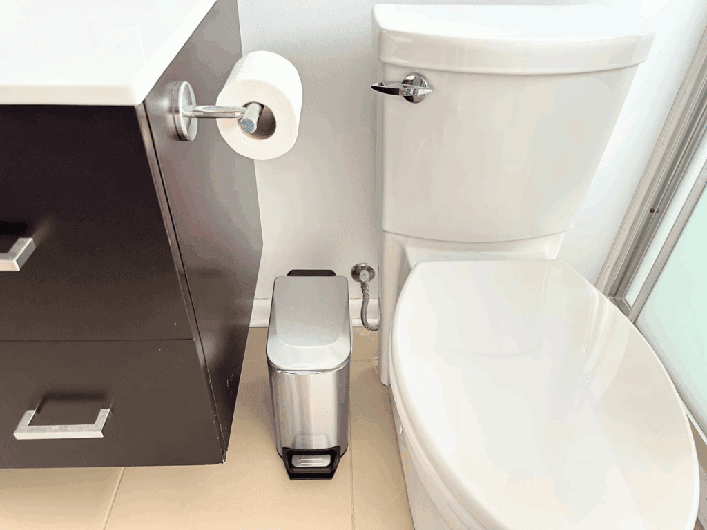 Bathroom corner with a white toilet next to a dark wood vanity. A chrome toilet paper holder with a roll is mounted on the side of the vanity. A small rectangular stainless steel step-on trash can is placed between the vanity and toilet. The floor is tiled in a light beige color.