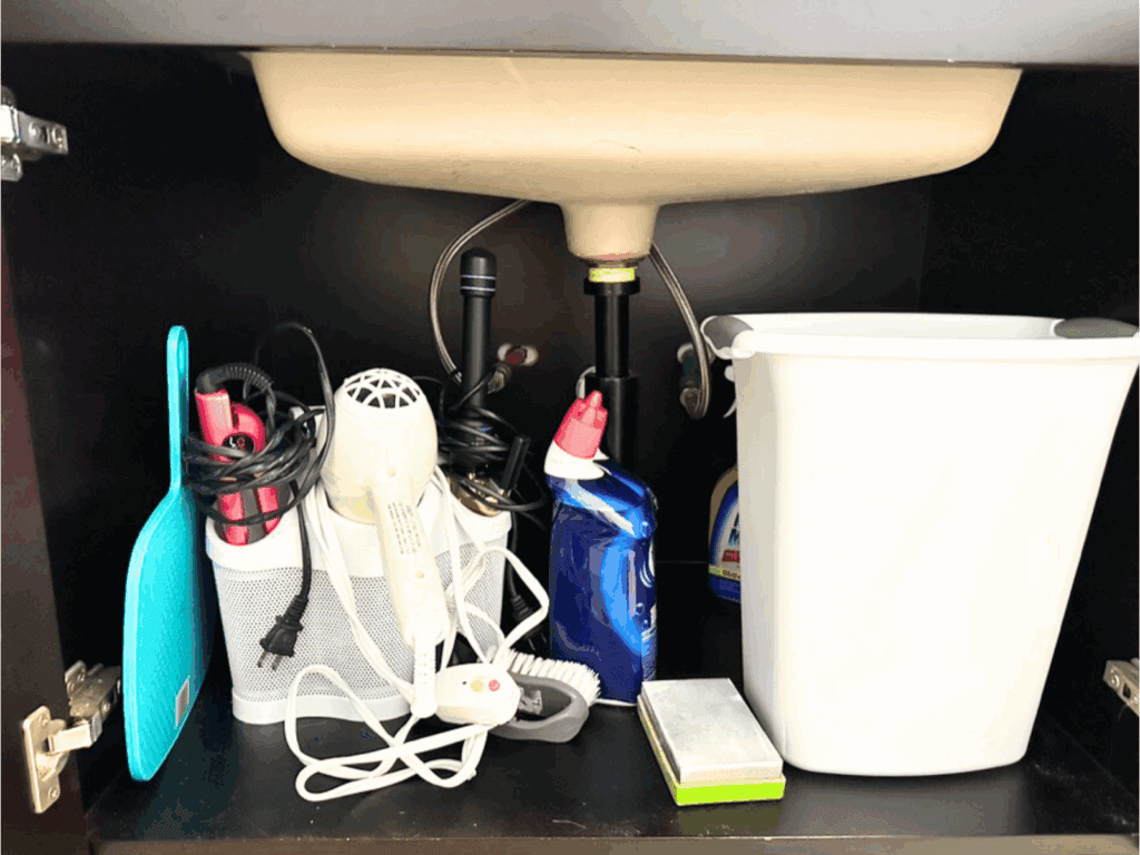 Under-sink bathroom cabinet with a white basin above. On the left, a turquoise mirror leans against a white mesh organizer holding a pink hair straightener, white hair dryer, and curling iron with cords wrapped around them. In the middle, a blue toilet cleaner bottle and small white cleaning brush sit next to a rectangular sponge. On the right, a tall white plastic trash can is placed beside a bottle of cleaning solution.