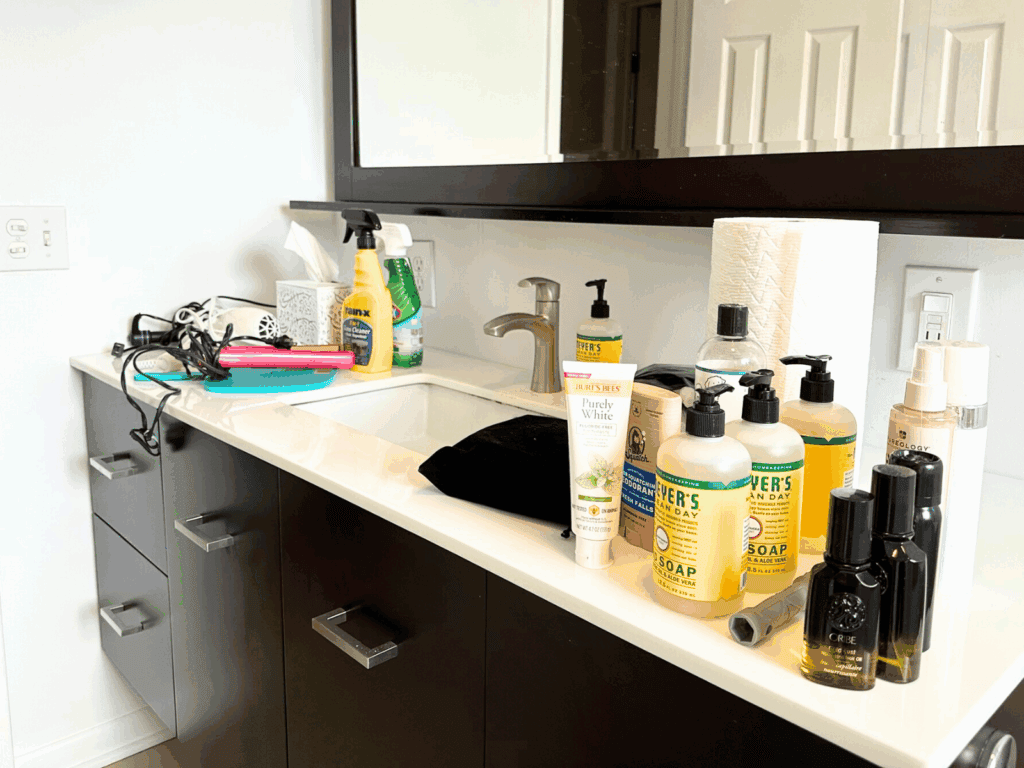 Bathroom countertop cluttered with items removed from under the sink. On the left side, hair styling tools including a curling iron, straightener, and hair dryer rest on top of a turquoise mirror near cleaning products and a tissue box. On the right side, multiple bottles of Mrs. Meyer’s hand soap, deodorant, toothpaste, hair products, a roll of paper towels, and other toiletries are grouped together near the sink.