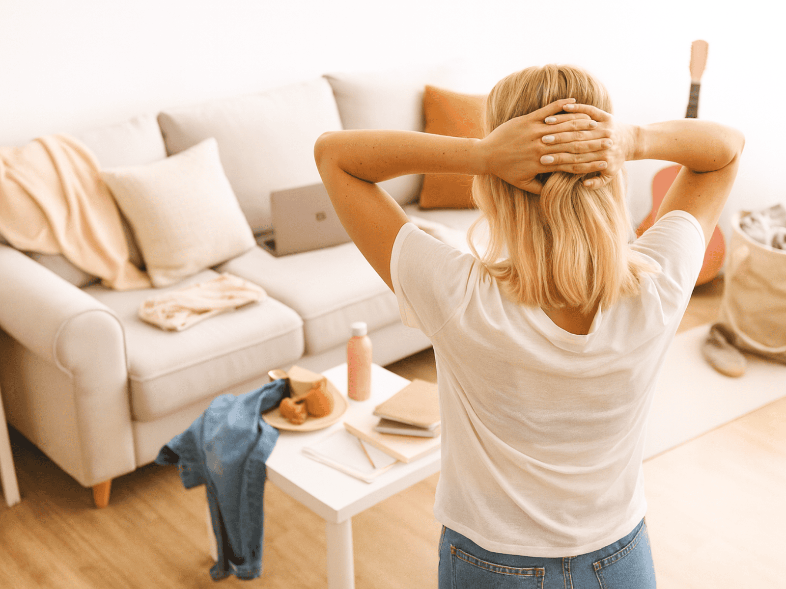 A blond woman in a white t-shirt and jeans stands with her hands on the back of her head, facing a cluttered living room. A white sofa with pastel and neutral decorative pillows is covered with scattered clothes and a blanket.