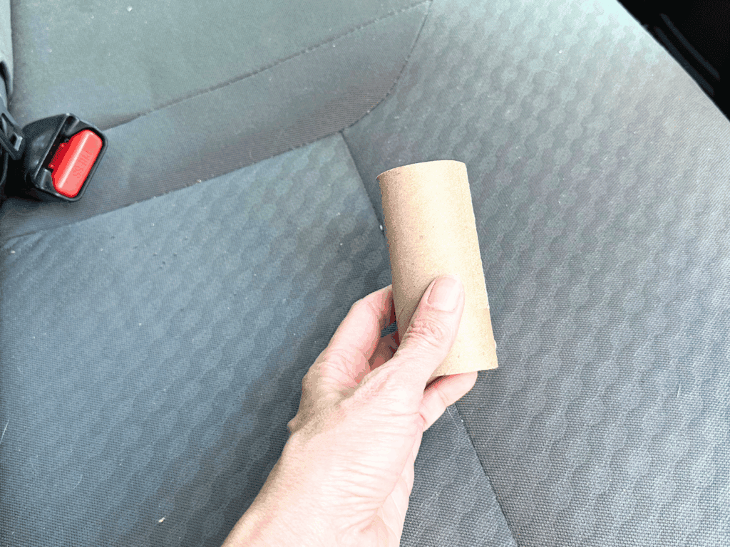 Hand holding an empty toilet paper roll on a car seat, used as a DIY air freshener by adding a few drops of essential oil.