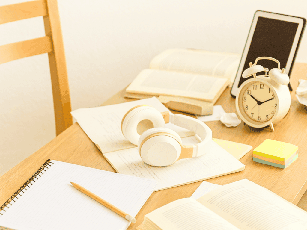 A slightly messy wooden desk with scattered notebooks, headphones, stationery, and small decor items, creating a realistic lived-in workspace scene.