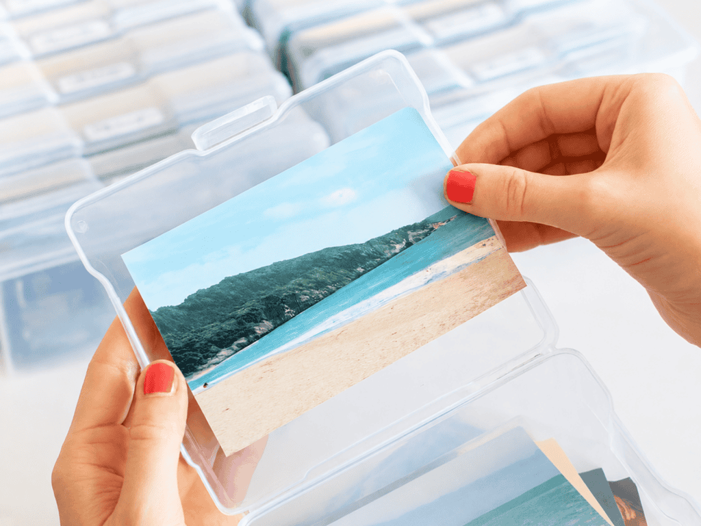 Hands sorting printed photographs into a clear plastic photo storage case, with multiple organized photo boxes in the background on a marble surface.