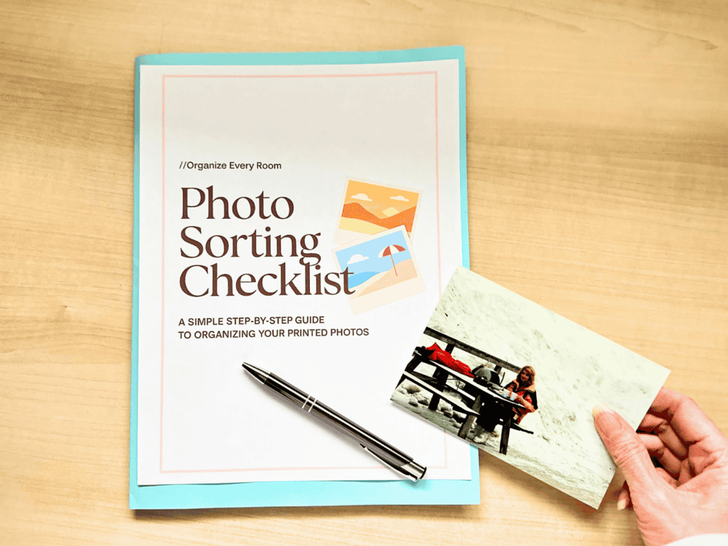 Printed photo sorting checklist on a light wood desk with a pen and a hand holding a vintage photograph, showing a simple and organized approach to sorting printed photos.