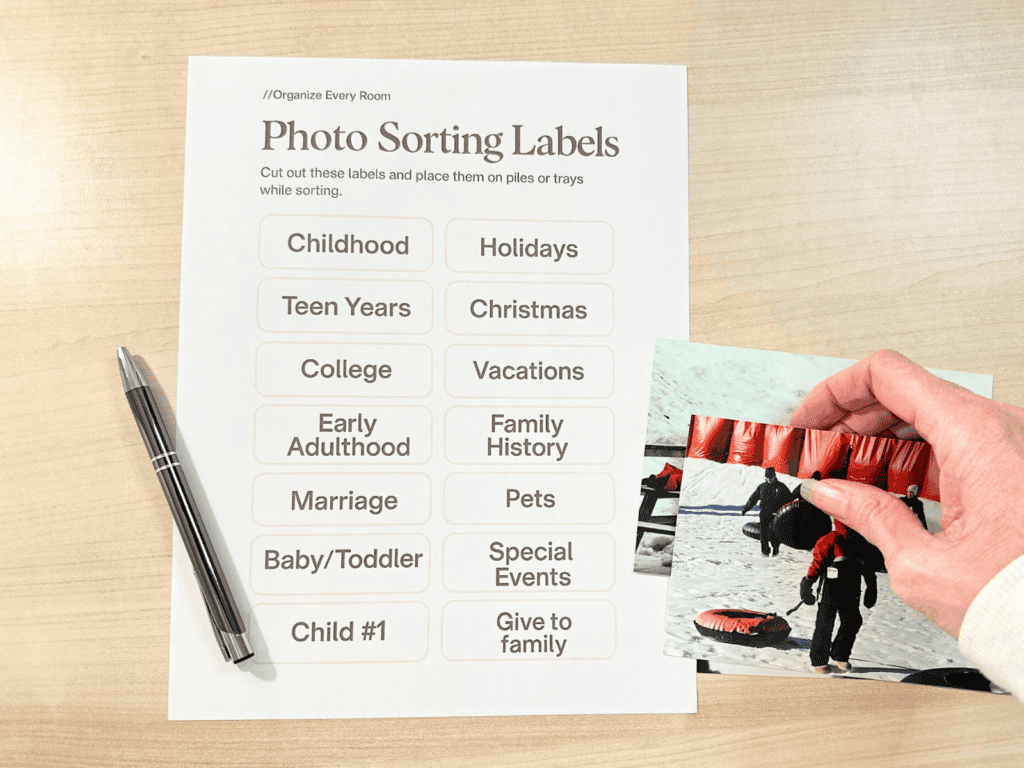 Flat lay of a photo sorting label sheet with categories like childhood, holidays, and vacations on a light wood desk, with a pen beside it and a hand holding printed photos.