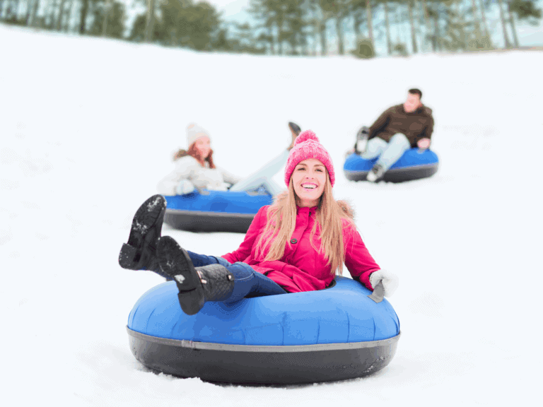 A smiling woman in a pink coat and pink winter hat rides a blue snow tube down a snowy hill, with two other people tubing in the background on a bright winter day.