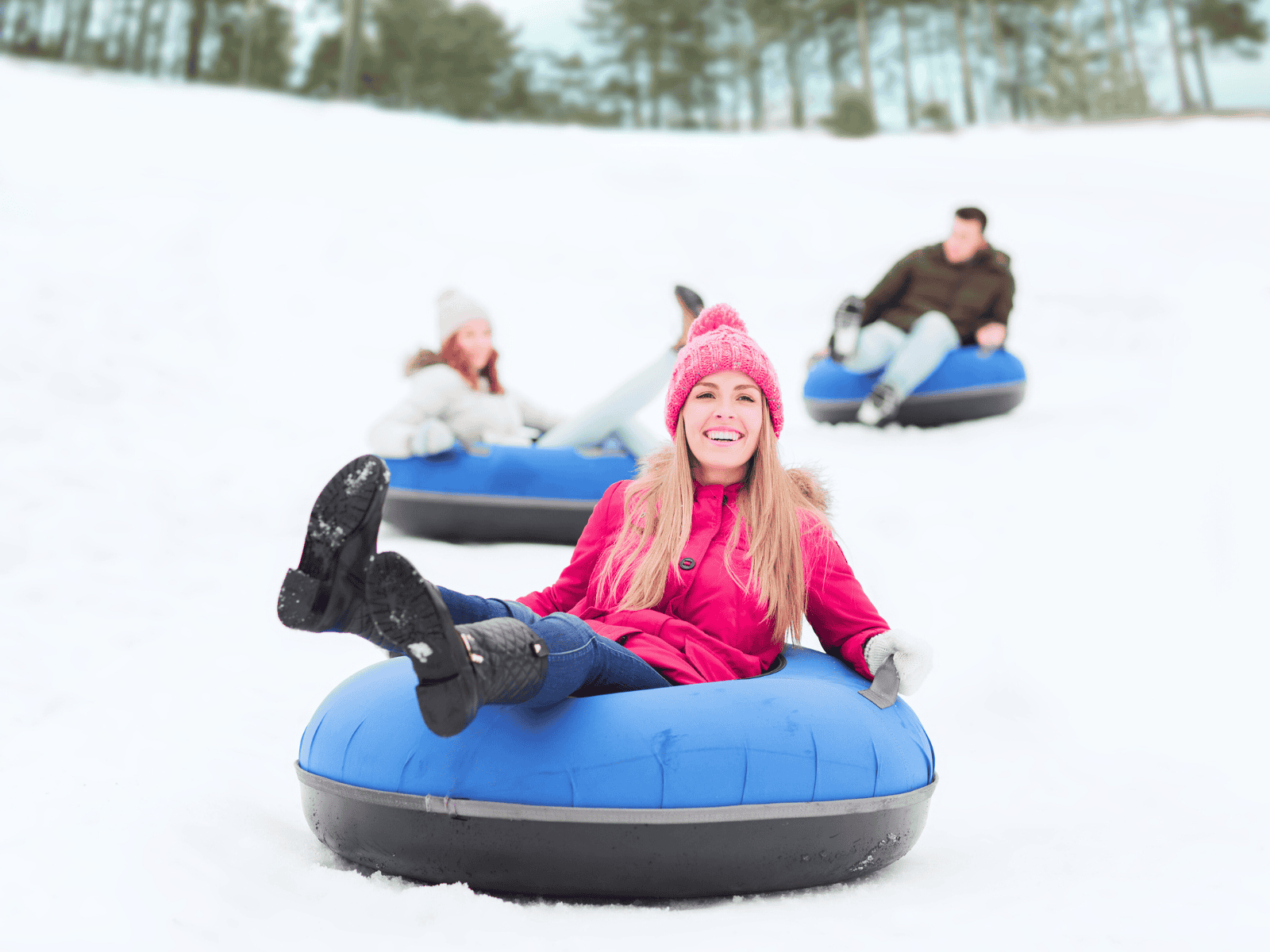A smiling woman in a pink coat and pink winter hat rides a blue snow tube down a snowy hill, with two other people tubing in the background on a bright winter day.