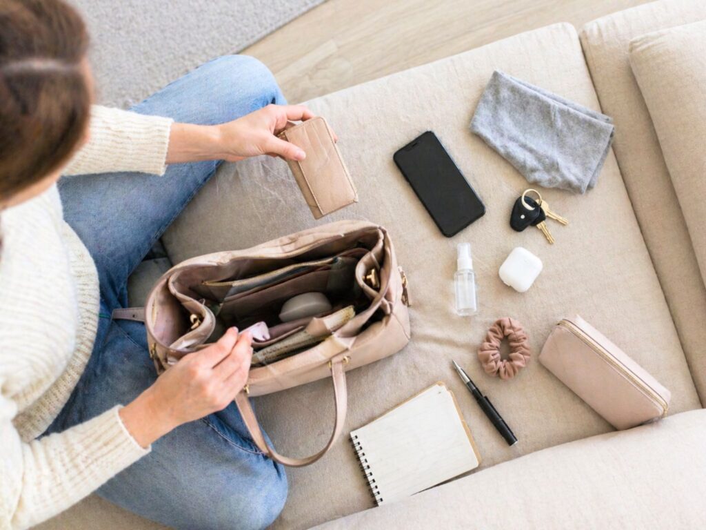 Top view of woman organizing handbag on beige sofa with wallet, phone, keys, notebook, pouch, and everyday essentials neatly spread out.